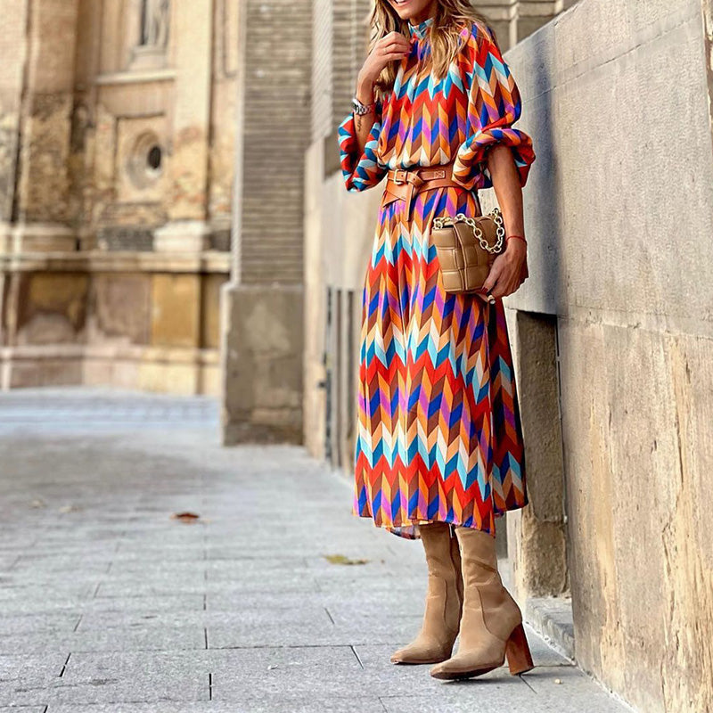 Frau in bunter Midi-Kleid mit geometrischem Muster, braunen Stiefeln und Tasche auf Straße.