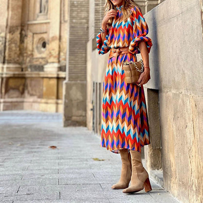 Frau in bunter Midi-Kleid mit geometrischem Muster, braunen Stiefeln und Tasche auf Straße.